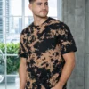 Man with short dark hair wearing a black and tan tie-dye T-shirt, standing with hands in pockets in a bright, modern indoor space.
