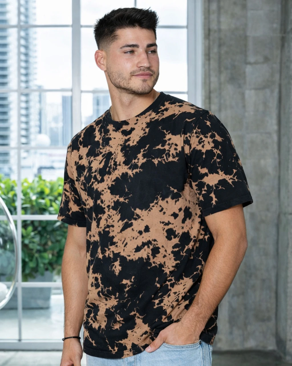 Man with short dark hair wearing a black and tan tie-dye T-shirt, standing with hands in pockets in a bright, modern indoor space.