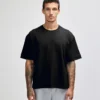 Man wearing a loose black T-shirt and light gray sweatpants stands facing the camera with a neutral expression, arms at sides, showing tattoo sleeves on both arms