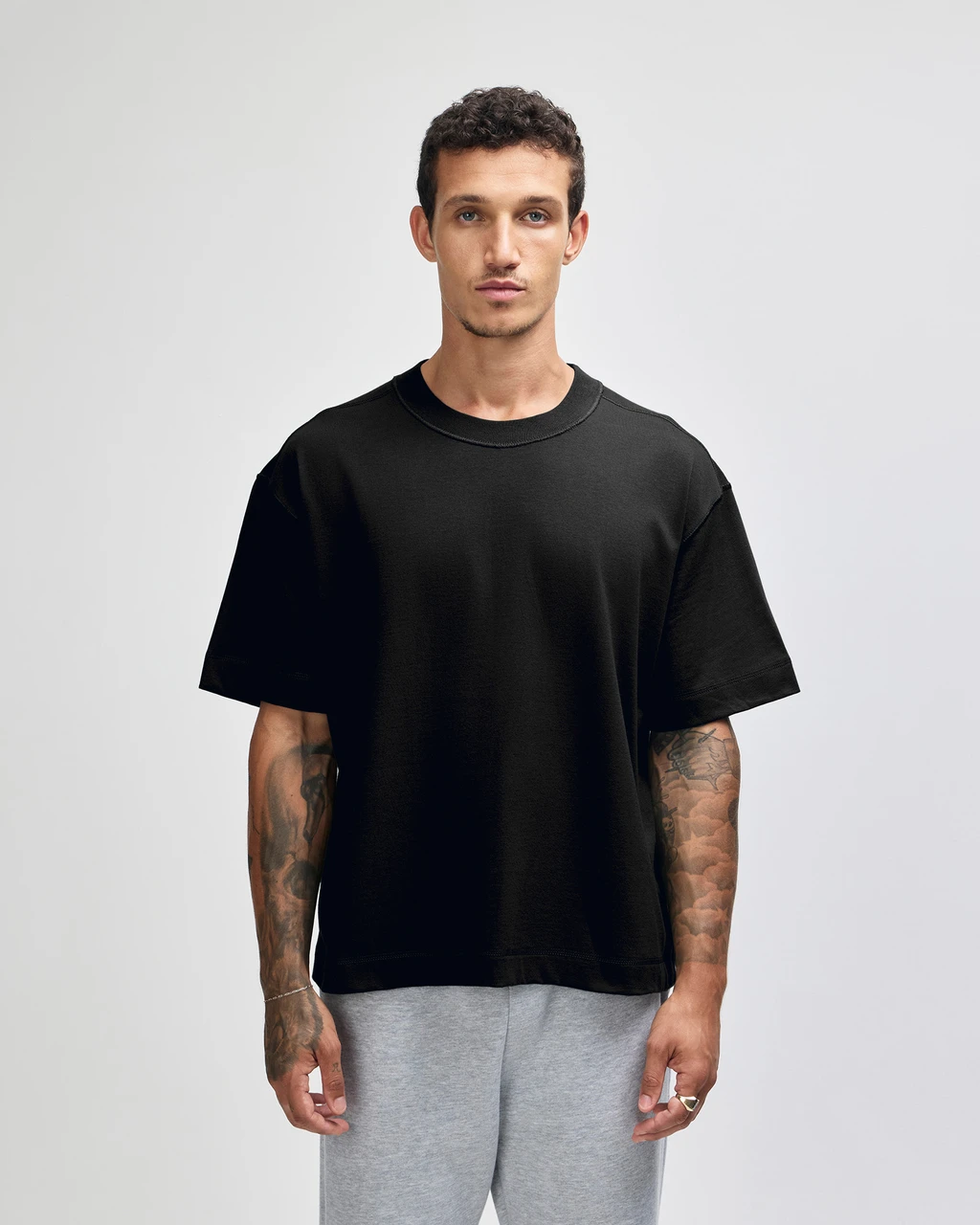 Man wearing a loose black T-shirt and light gray sweatpants stands facing the camera with a neutral expression, arms at sides, showing tattoo sleeves on both arms