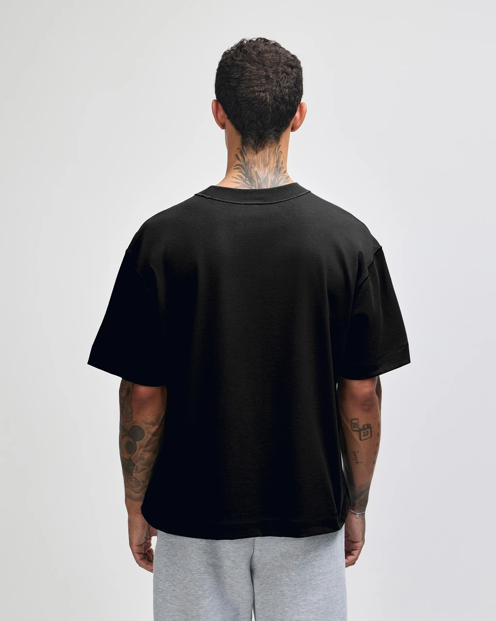 Back view of a person wearing a black oversized T-shirt and gray shorts, with tattoos on the neck and arms visible.