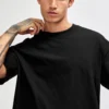Man wearing a plain black crewneck T-shirt, posing with hand near the collar.