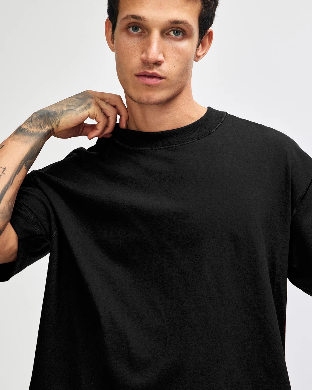 Man wearing a plain black crewneck T-shirt, posing with hand near the collar.