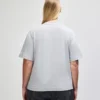Back view of a person wearing a light gray T-shirt and dark jeans, with long blonde hair tied back, standing against a plain light background.
