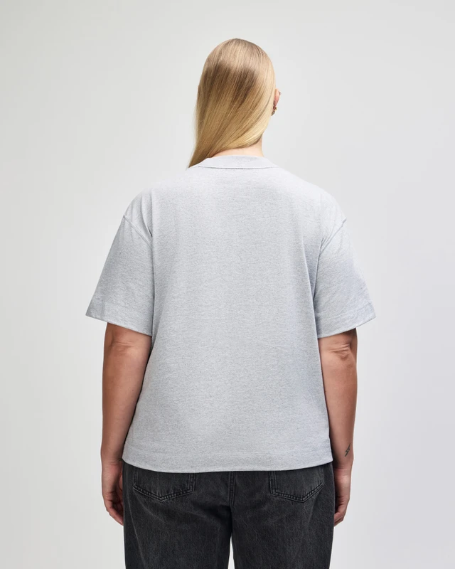Back view of a person wearing a light gray T-shirt and dark jeans, with long blonde hair tied back, standing against a plain light background.