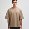 Man with short dark hair and tattoos wearing a beige oversized short-sleeve sweatshirt, standing against a light background with a neutral expression.