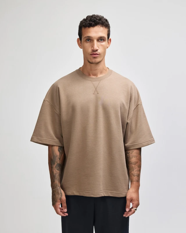 Man with short dark hair and tattoos wearing a beige oversized short-sleeve sweatshirt, standing against a light background with a neutral expression.