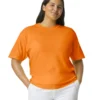 Woman wearing a bright orange T-shirt with white pants, standing with hands in pockets and a slight smile against a white background.