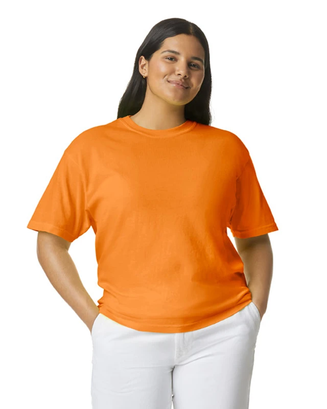 Woman wearing a bright orange T-shirt with white pants, standing with hands in pockets and a slight smile against a white background.