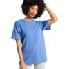 Woman with curly hair wearing a blue T-shirt and light pants, posing with one hand on her head against a white background.