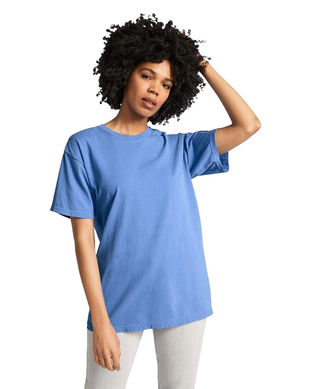Woman with curly hair wearing a blue T-shirt and light pants, posing with one hand on her head against a white background.