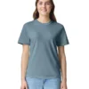 Woman standing and smiling, wearing a blue-gray T-shirt and light jeans, facing the camera against a white background.