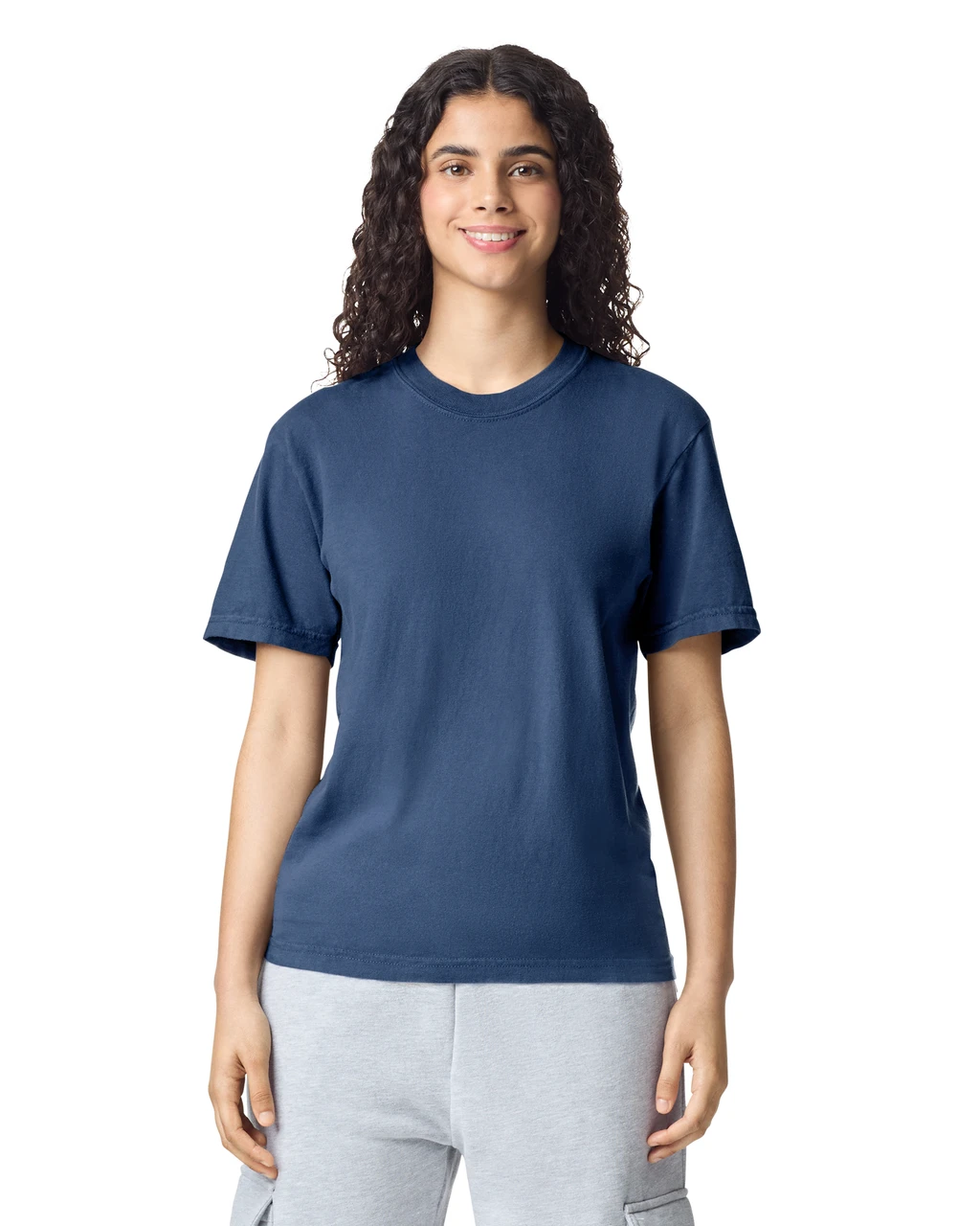 Woman with dark curly hair wearing a navy T-shirt and light gray joggers, standing against a white background, smiling at the camera.