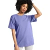Woman with curly hair wearing a loose lavender T-shirt and light gray pants, one hand running through her hair in a casual pose.