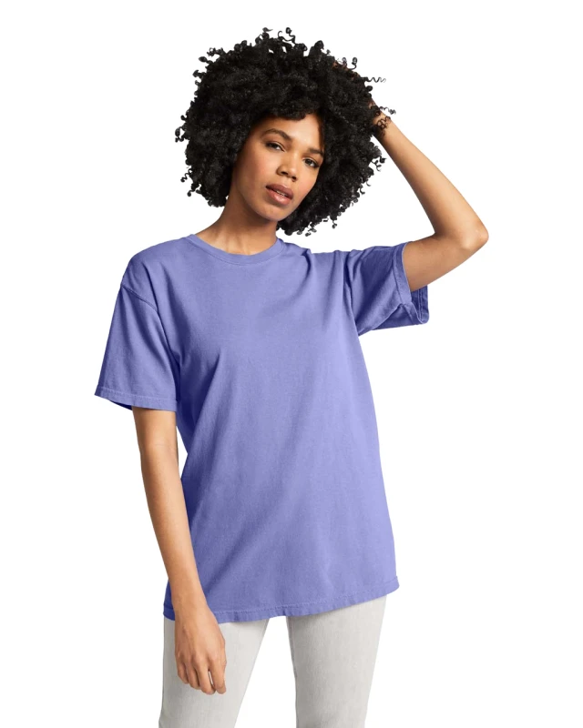 Woman with curly hair wearing a loose lavender T-shirt and light gray pants, one hand running through her hair in a casual pose.