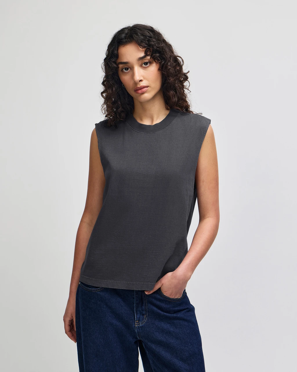 207GD_FadedBlack_05__74348 Woman modeling a dark gray sleeveless top with blue jeans, standing with one hand in her pocket against a light backdrop.