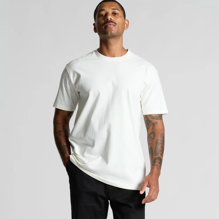 5026G_CLASSIC_ORGANIC_TEE_LOOSE__60124 Man wearing a plain white crew-neck T-shirt and black pants, standing against a light gray background.