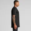 5070_CLASSIC_PLUS_TEE_SIDE__11966 Side profile of a man wearing a black short-sleeve T-shirt, with a tattooed arm, against a gray studio backdrop.