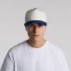 CLASS_2_TONE_TURN__13362 Young man wearing a white baseball cap with a blue brim and a plain white T‑shirt, facing the camera against a light gray background.