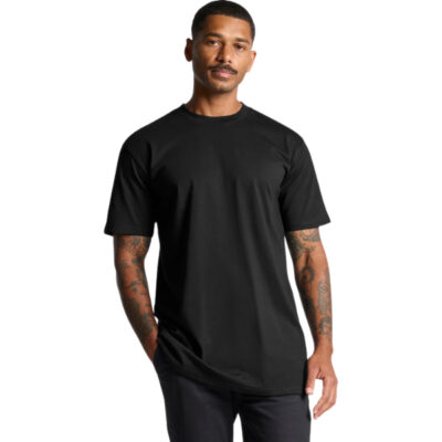 Man in a black short-sleeve T-shirt standing with one hand in his pocket, tattoos visible on his arms.